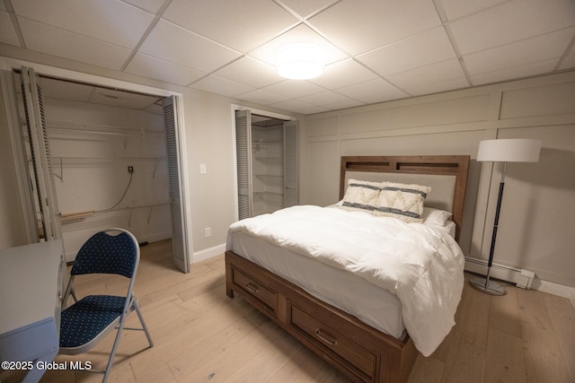 bedroom featuring multiple closets, light hardwood / wood-style flooring, a baseboard radiator, and a drop ceiling