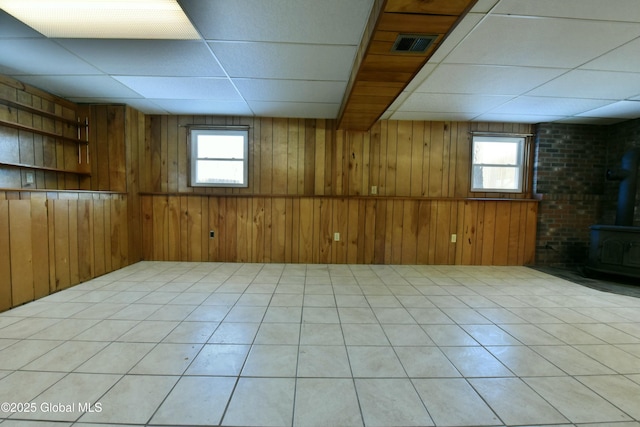 basement with light tile patterned floors, a wood stove, a drop ceiling, and wooden walls