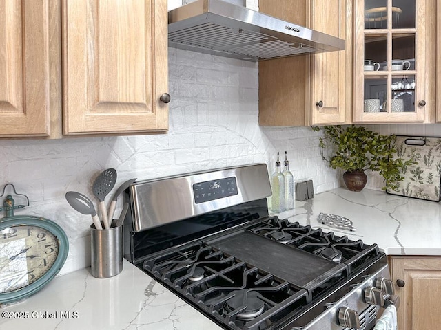 kitchen with range hood, stainless steel gas range oven, light stone countertops, light brown cabinetry, and tasteful backsplash