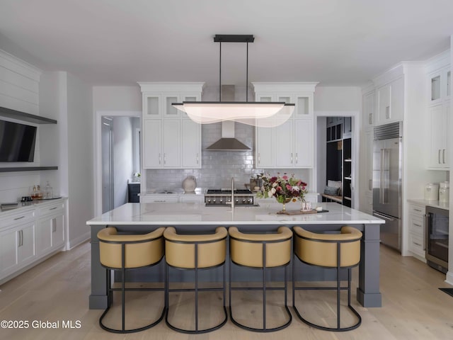 kitchen featuring wall chimney range hood, built in fridge, decorative light fixtures, and a center island with sink