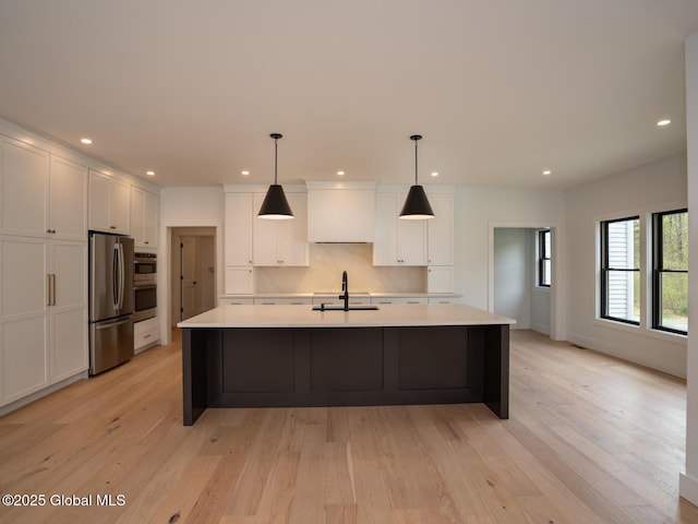 kitchen with white cabinets, light wood-type flooring, a center island with sink, pendant lighting, and appliances with stainless steel finishes