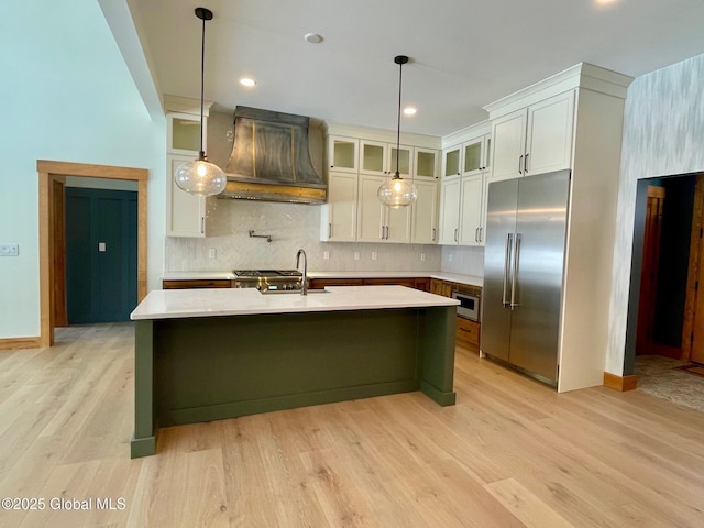 kitchen with light countertops, appliances with stainless steel finishes, glass insert cabinets, an island with sink, and premium range hood