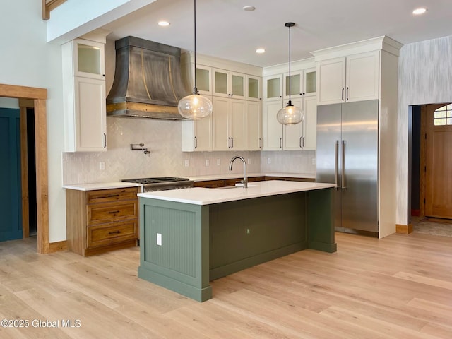 kitchen featuring premium range hood, light countertops, appliances with stainless steel finishes, an island with sink, and glass insert cabinets