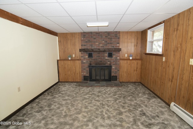 unfurnished living room with baseboard heating, wooden walls, dark carpet, a fireplace, and a drop ceiling