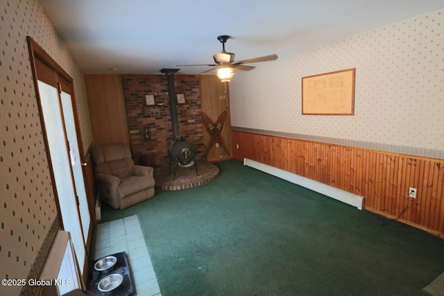 unfurnished room featuring wood walls, ceiling fan, a wood stove, carpet floors, and baseboard heating