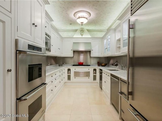 kitchen featuring wall chimney range hood, white cabinetry, a sink, and built in appliances