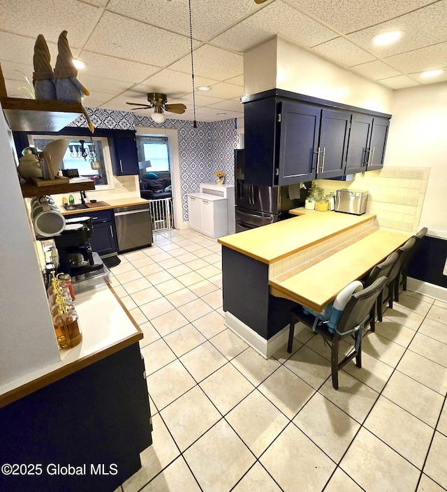 kitchen featuring blue cabinets, light tile patterned floors, stainless steel dishwasher, ceiling fan, and a drop ceiling