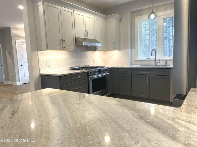 kitchen featuring decorative backsplash, light stone countertops, high end stove, under cabinet range hood, and a sink