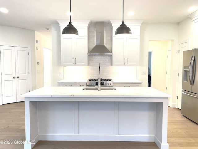 kitchen with wall chimney exhaust hood, light stone counters, stainless steel fridge with ice dispenser, hanging light fixtures, and a center island with sink