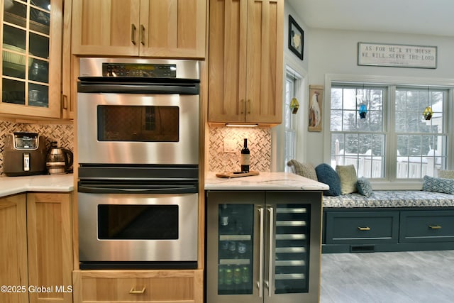 kitchen with beverage cooler, decorative backsplash, light stone counters, glass insert cabinets, and stainless steel double oven