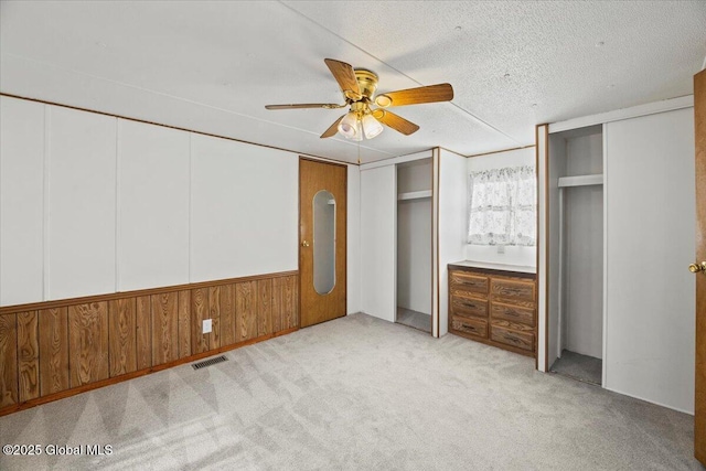 unfurnished bedroom with wood walls, light colored carpet, a textured ceiling, two closets, and ceiling fan