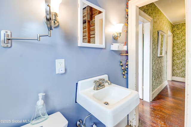 bathroom featuring wallpapered walls, baseboards, a sink, and wood finished floors