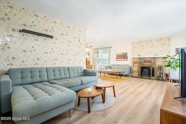 living room featuring wallpapered walls, a ceiling fan, wood finished floors, and a stone fireplace