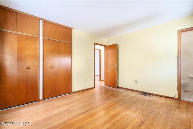 unfurnished bedroom featuring ensuite bath, light wood finished floors, baseboards, and two closets