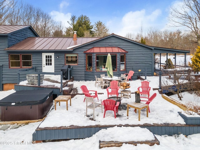 snow covered back of property with metal roof, an outdoor fire pit, a standing seam roof, a patio area, and a hot tub