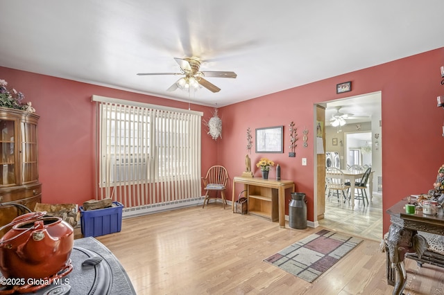 living area with ceiling fan, a baseboard heating unit, wood finished floors, baseboards, and washer / dryer