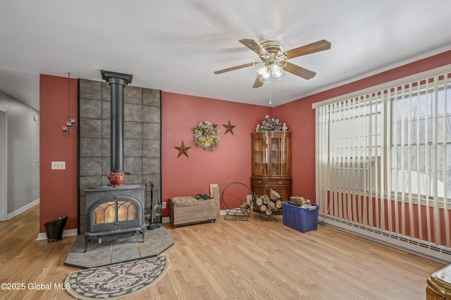 sitting room with baseboards, a ceiling fan, wood finished floors, a wood stove, and a baseboard heating unit