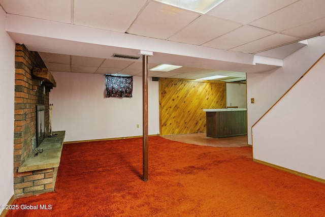 finished basement with wooden walls, visible vents, a drop ceiling, carpet flooring, and a fireplace