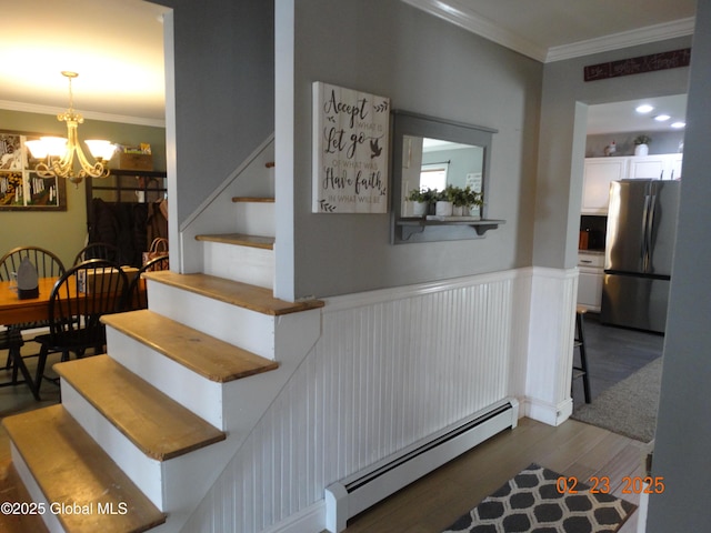 stairway with wainscoting, ornamental molding, wood finished floors, baseboard heating, and a chandelier