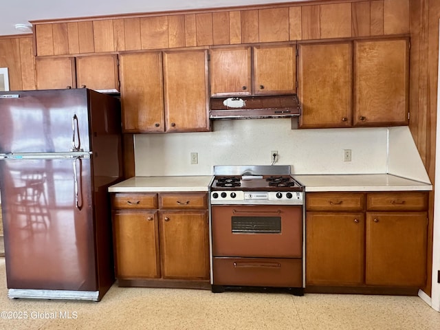 kitchen with brown cabinetry, freestanding refrigerator, light countertops, under cabinet range hood, and range with gas cooktop