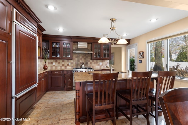 kitchen featuring premium appliances, backsplash, glass insert cabinets, a sink, and wall chimney exhaust hood