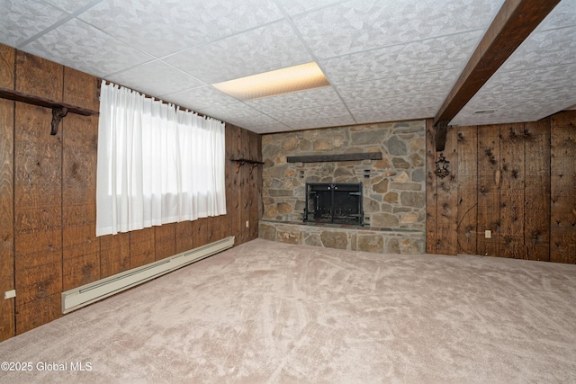 unfurnished living room featuring a stone fireplace, a paneled ceiling, wooden walls, a baseboard heating unit, and carpet