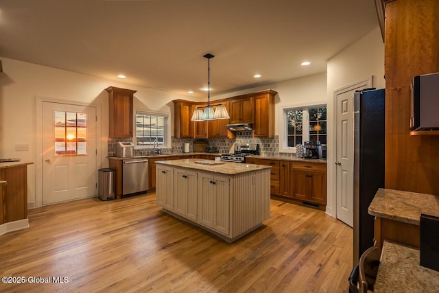 kitchen featuring stainless steel appliances, tasteful backsplash, light wood-style flooring, brown cabinetry, and under cabinet range hood