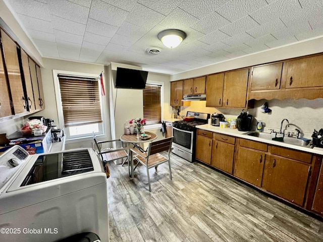 kitchen featuring brown cabinetry, a sink, washer / dryer, under cabinet range hood, and stainless steel gas range oven