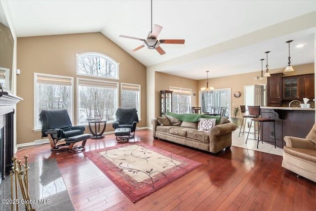 living room with hardwood / wood-style floors, a glass covered fireplace, high vaulted ceiling, baseboards, and ceiling fan with notable chandelier