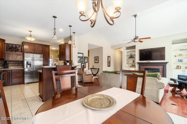 dining space with built in shelves, a fireplace, light tile patterned floors, lofted ceiling, and ceiling fan with notable chandelier