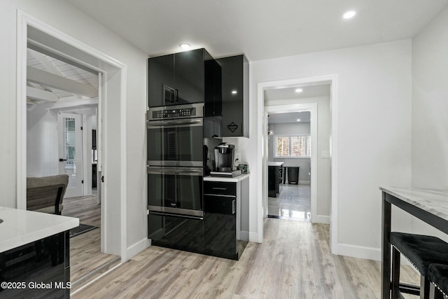 kitchen with stainless steel double oven, baseboards, dark cabinetry, light wood-type flooring, and light stone countertops