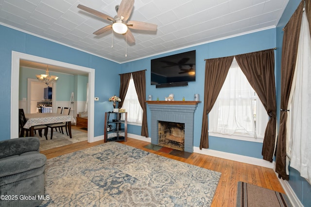 living room with a wealth of natural light, a brick fireplace, wood finished floors, and ceiling fan with notable chandelier