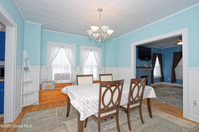 dining space with a wainscoted wall, ornamental molding, ceiling fan with notable chandelier, a fireplace, and wood finished floors