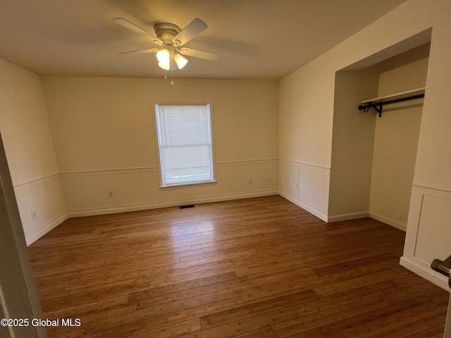 unfurnished bedroom featuring visible vents, a ceiling fan, wainscoting, wood-type flooring, and a decorative wall