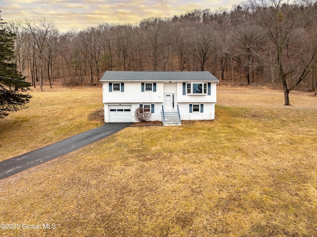 raised ranch with a garage, a forest view, a front lawn, and aphalt driveway