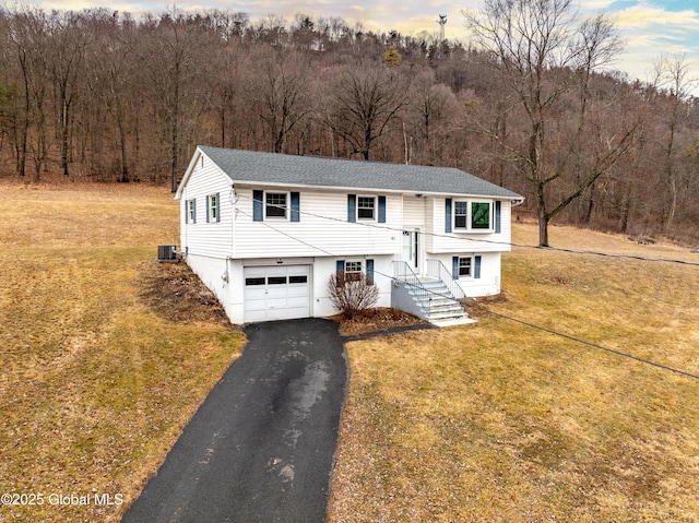 split foyer home featuring aphalt driveway, an attached garage, cooling unit, a front yard, and a view of trees