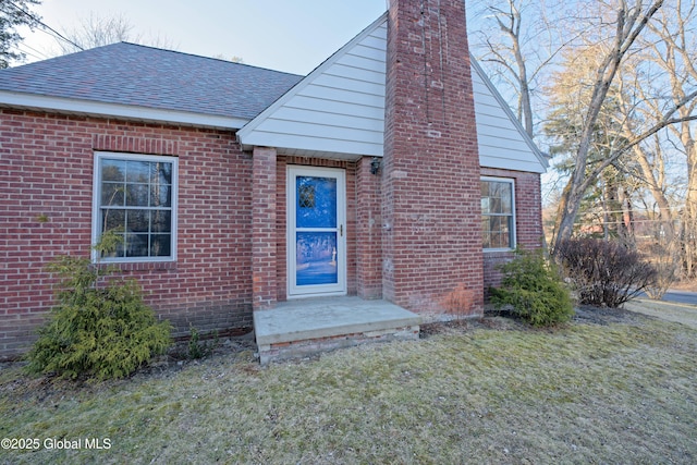 doorway to property with a yard, brick siding, roof with shingles, and a chimney