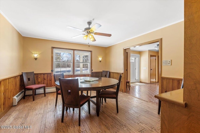 dining area featuring wooden walls, a ceiling fan, a wainscoted wall, light wood-style floors, and a baseboard heating unit