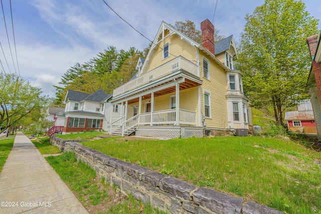 victorian-style house featuring a chimney, a porch, a balcony, cooling unit, and a front lawn