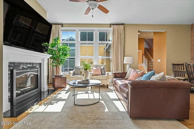 living room featuring a glass covered fireplace, stairs, ceiling fan, and wood finished floors