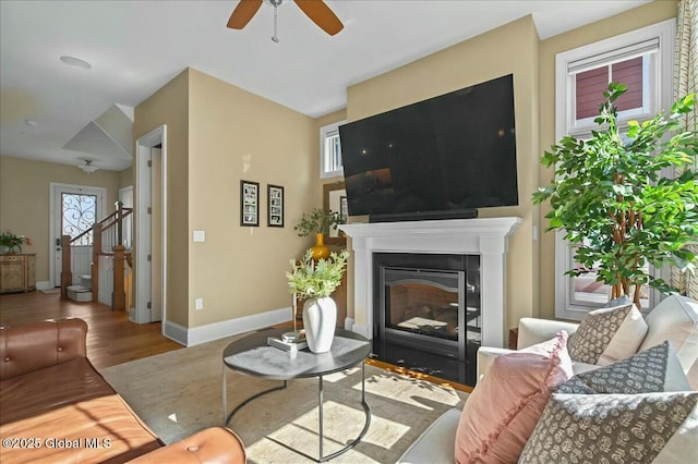 living area with a fireplace with flush hearth, wood finished floors, stairway, baseboards, and ceiling fan