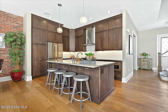 kitchen featuring light wood-style floors, dark brown cabinets, freestanding refrigerator, and wall chimney range hood