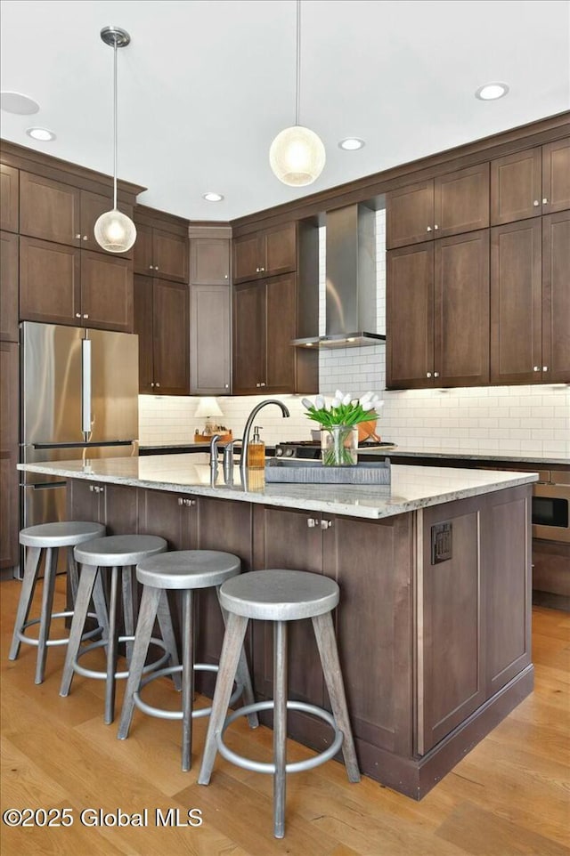 kitchen featuring freestanding refrigerator, dark brown cabinets, wall chimney exhaust hood, and light wood-style floors