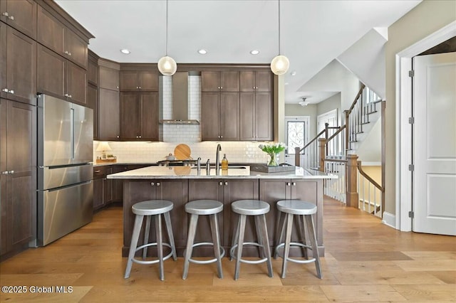 kitchen featuring light wood finished floors, dark brown cabinetry, wall chimney exhaust hood, and freestanding refrigerator