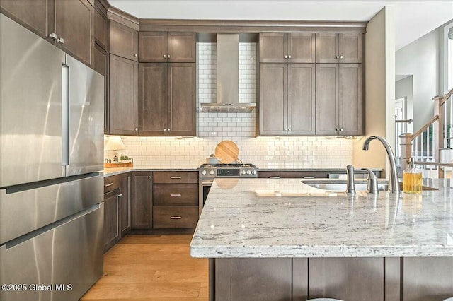 kitchen with backsplash, dark brown cabinetry, light wood-type flooring, stainless steel appliances, and wall chimney exhaust hood