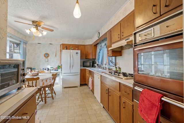 kitchen with wallpapered walls, under cabinet range hood, light countertops, brown cabinets, and white appliances