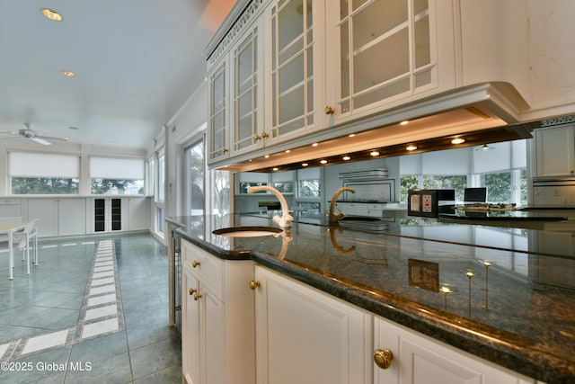 kitchen featuring tile patterned flooring, glass insert cabinets, ceiling fan, dark stone countertops, and a sink