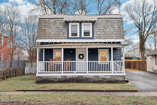 view of front of house with covered porch, a gambrel roof, roof with shingles, and fence
