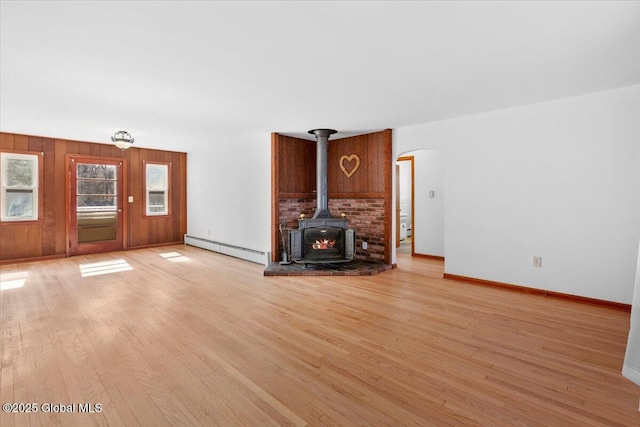 unfurnished living room featuring a baseboard heating unit, wooden walls, light wood finished floors, baseboards, and a wood stove