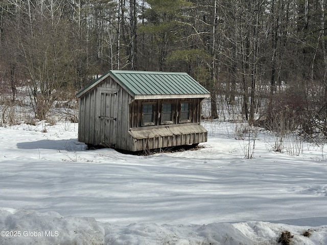 snow covered structure with an outdoor structure, a wooded view, and a shed